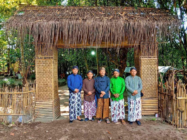 Five men in traditional clothing pose under a thatched wooden gate in a rural village setting